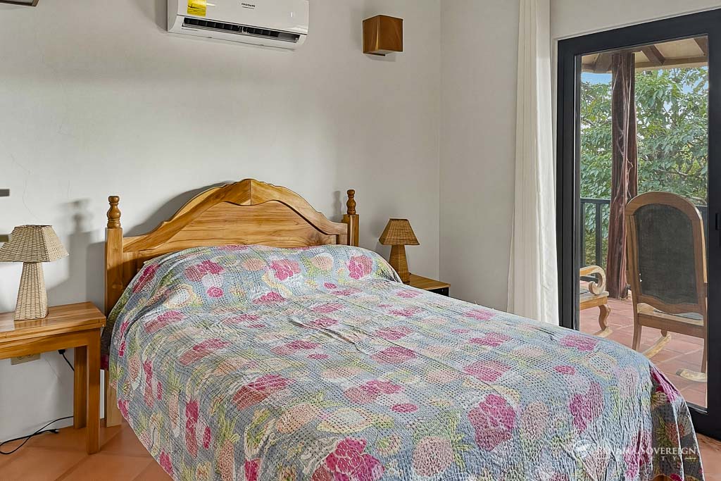 Habitación con cama de madera, colcha floral, mesas de noche y acceso a balcón con vista verde