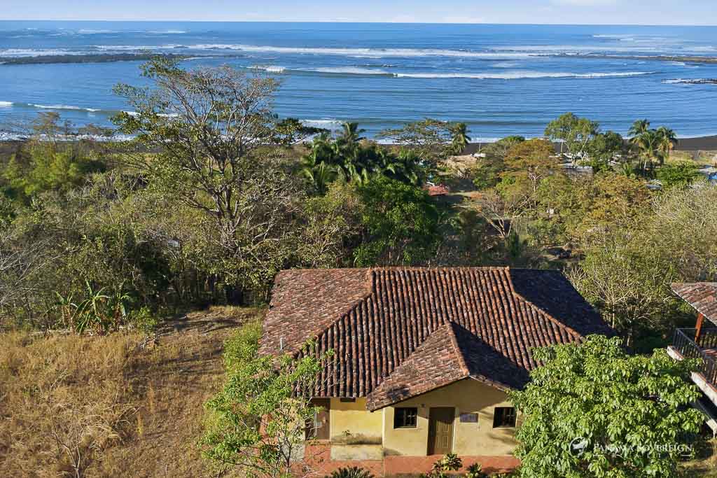 Vista aérea de casa con techo de tejas rodeada de vegetación y olas del océano al fondo