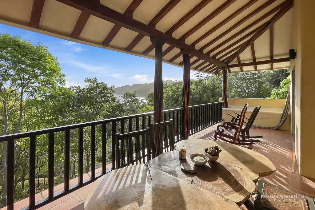 Terraza techada con mecedoras, mesa de madera y vista hacia vegetación y costa