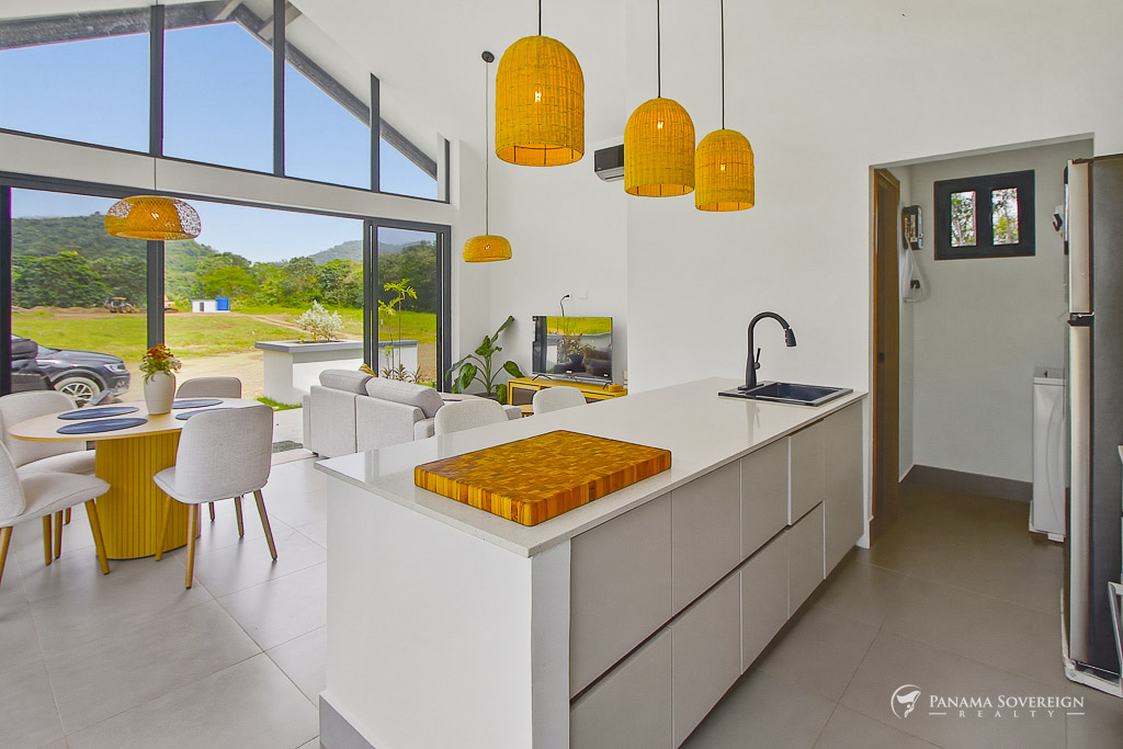 Espacio abierto de cocina y sala con techos altos, isla central, lámparas colgantes y grandes ventanales con vista al exterior