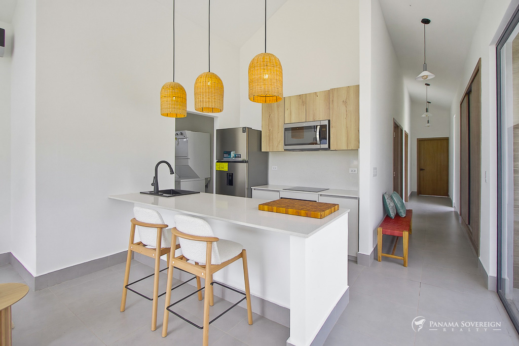 Cocina moderna con isla blanca, dos taburetes, electrodomésticos de acero inoxidable y pasillo hacia habitaciones