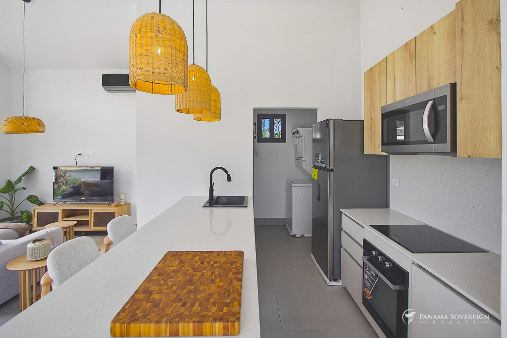 Cocina equipada con encimera blanca, horno empotrado, microondas, refrigerador y vista hacia la sala