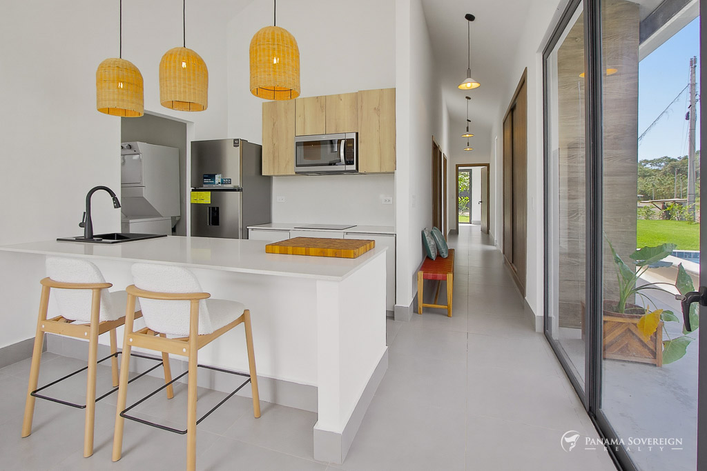 Cocina abierta y luminosa con isla blanca, gabinetes de madera, lámparas colgantes, electrodomésticos de acero y vista al pasillo
