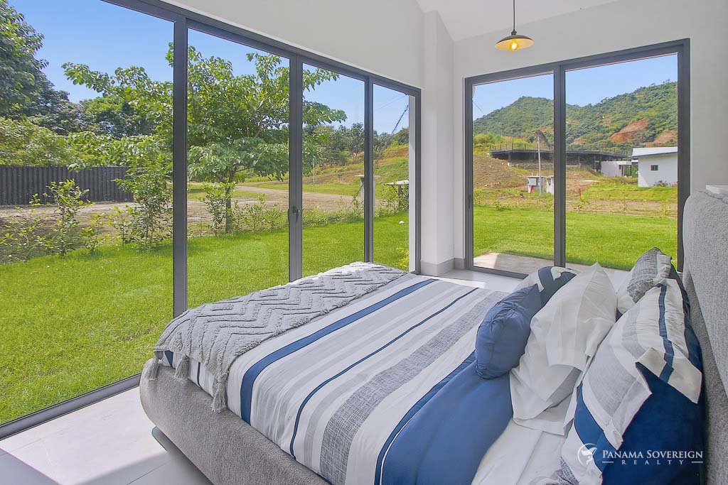 Dormitorio con ventanales de piso a techo, vista a jardín verde y colinas, cama moderna y decoración en tonos neutros