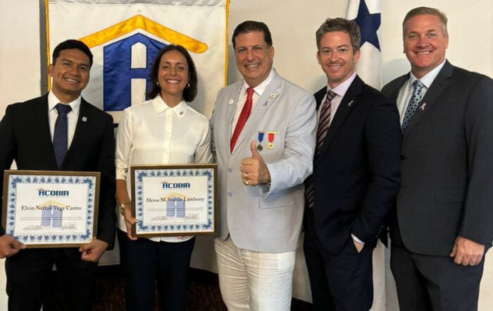 The image shows a group of five individuals posing together. Two of them, Elvin Nefatal Vega Castro and Alessa M. Stabile Lamberty, are holding certificates from ACOBIR. They are being congratulated by Alvaro Boyd who is giving a thumbs up. The group is standing in front of an ACOBIR banner, and a Panamanian flag is visible in the background. This is an induction ceremony for the two new ACOBIR members.