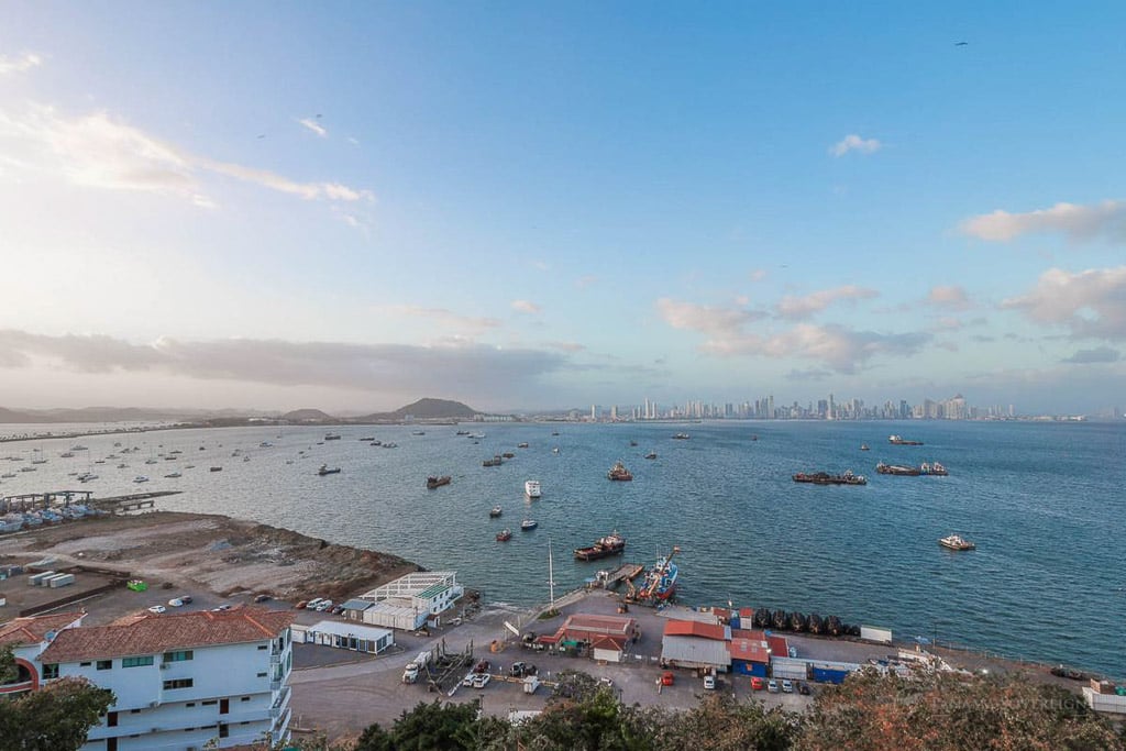 A wide view of the coastline, with ships in the water, a distant city skyline, and a clear blue sky. In the foreground, red-roofed buildings and industrial structures are visible near the water.