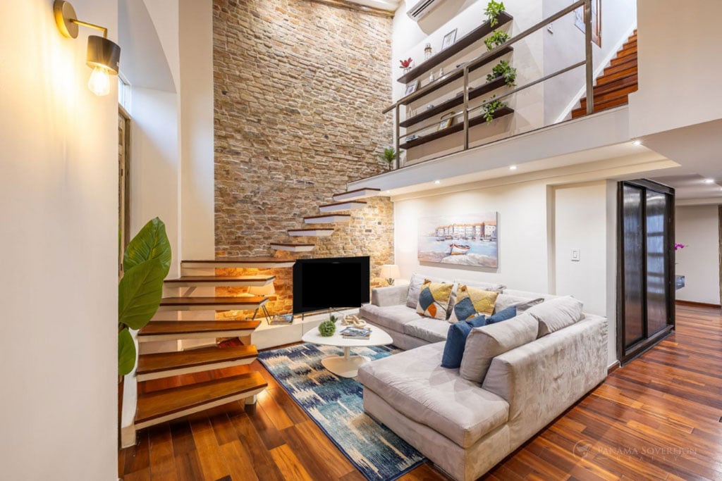 Cozy living area with an exposed brick wall, floating wooden staircase, sectional couch, and bright natural light from large windows.