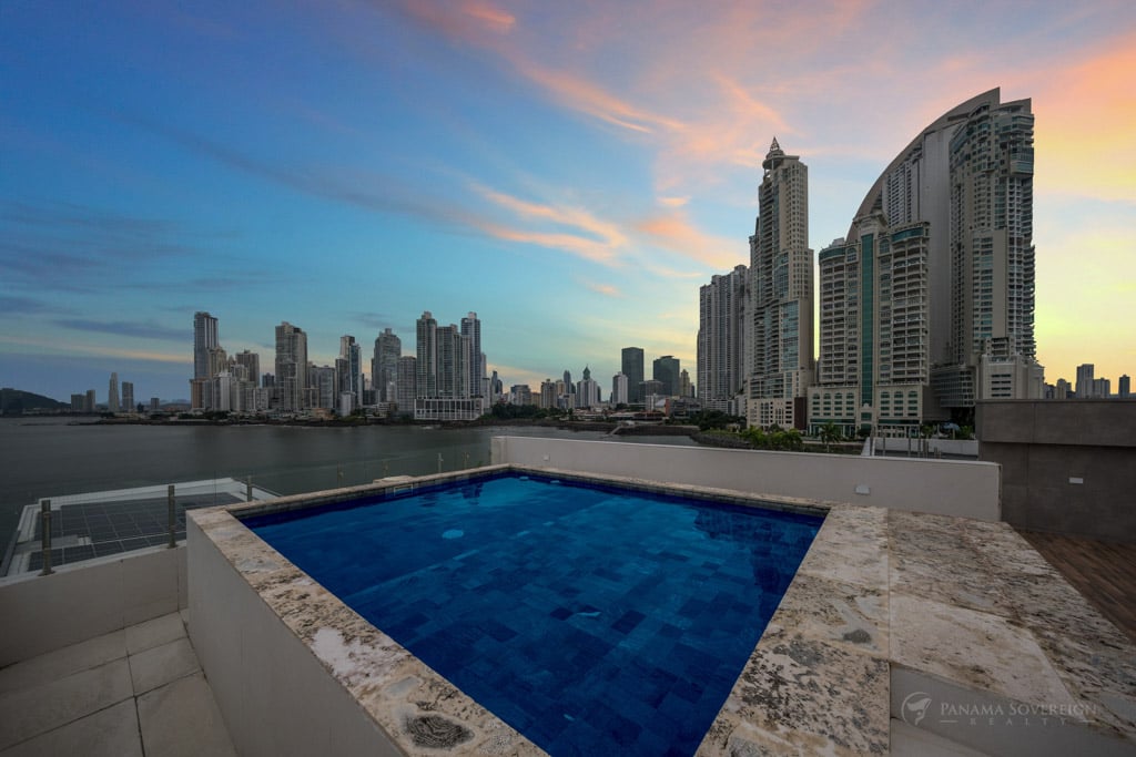 A rooftop pool with deep blue tiles, overlooking Panama City’s skyline and waterfront. The sunset casts a warm glow on the cityscape.