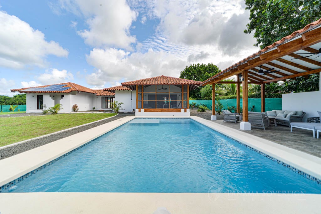 A large, inviting swimming pool in the backyard of a modern, single-story white house with a red-tiled roof and wooden beams.