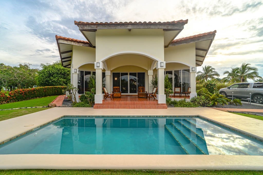 The front view of the house with a large covered patio, terra cotta tiles, and a private pool in the foreground. The house’s white facade contrasts beautifully with the lush green lawn and tropical plants surrounding the area.