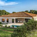 Side angle of Pedasí home with pool, covered terrace, lush bushes, and palm trees.
