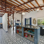 Open-plan kitchen and living area with center island, vaulted ceilings, exposed beams, and decorative tile throughout.