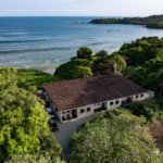 Ocean-facing aerial of triplex structure with red tile roof, located just meters from the waterline with lush trees and gentle surf below.