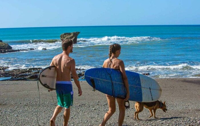 A man and woman walking along the beach in Cambutal, Panama, carrying surfboards with their dog walking beside them.