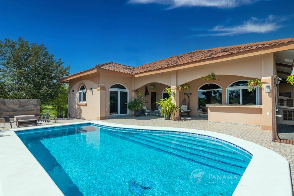 Vista junto a la piscina de una casa de lujo con tejado de tejas rojas, que muestra una piscina azul inmaculada y una zona de patio.