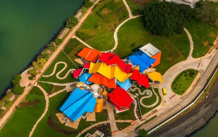 Aerial view of the Museo de la Biodiversidad, highlighting its brightly colored, geometric roof design. The museum is surrounded by green lawns and pathways, with the ocean visible nearby.