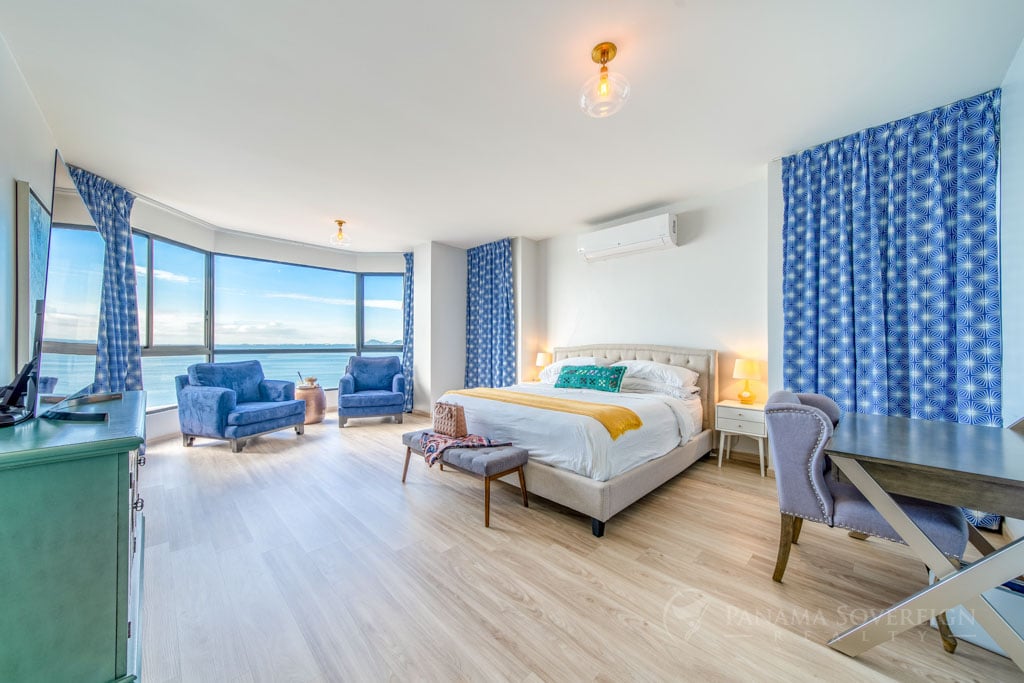 Spacious bedroom with large windows offering ocean views and blue armchairs.