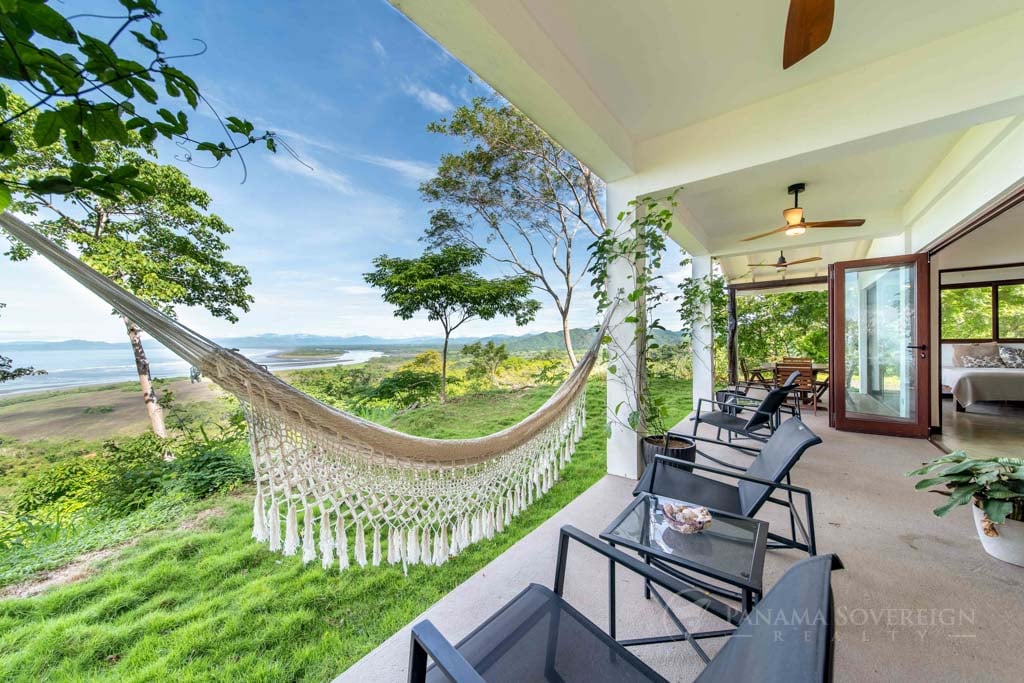 Covered outdoor patio with a hammock and seating, overlooking a lush green lawn and expansive ocean views.