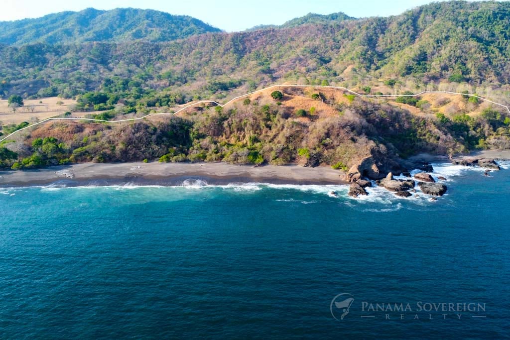 Aerial view of the coastline showing the marked property extending along the hills. The image captures the waves crashing onto a dark sandy beach, with the rugged terrain and hills covered in a mix of trees and dry grass.