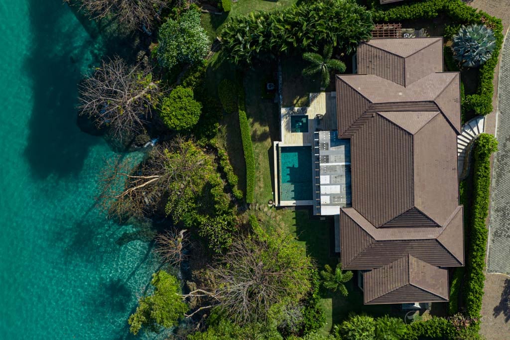 Aerial view of a luxury villa with a private pool, situated on a waterfront property surrounded by lush greenery, with the turquoise waters of the ocean just beyond the tree line.