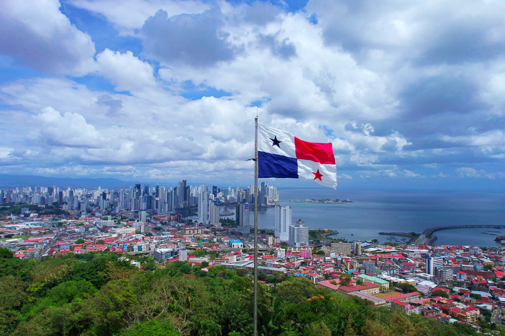 Panamanian flag waving on a hill with a sweeping view of Panama City’s skyline and the Pacific Ocean under a cloudy sky.