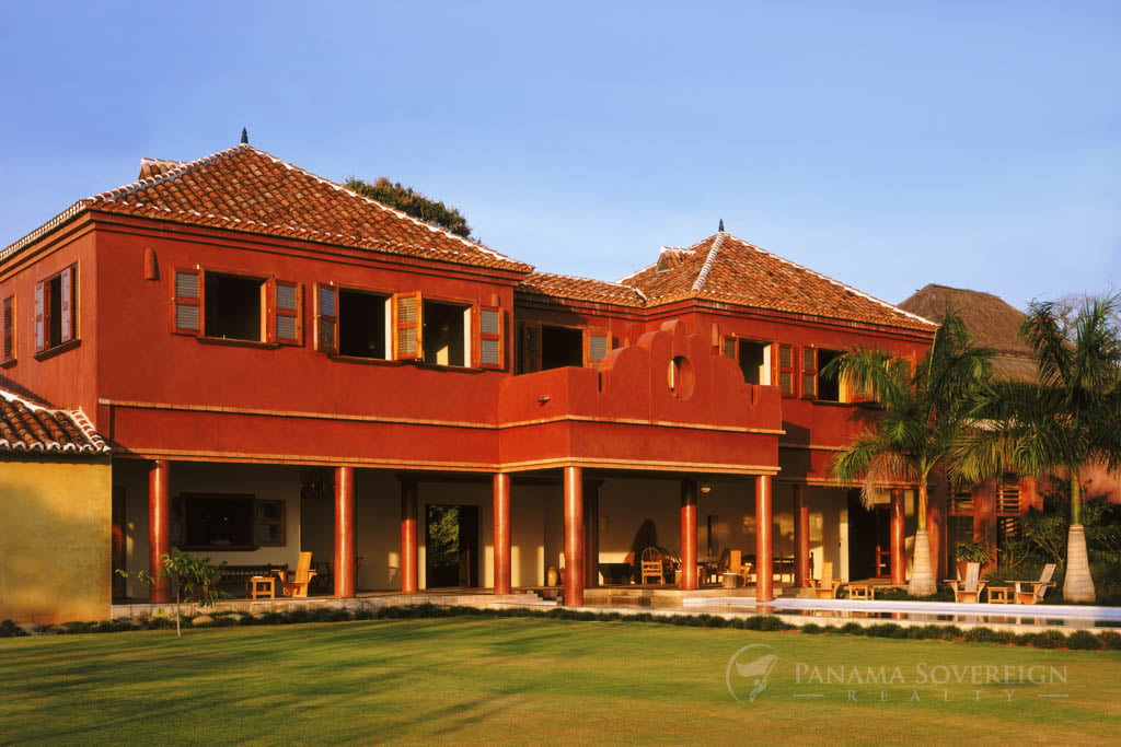 A vibrant, terracotta-colored two-story house with wooden shutters and a tiled roof, surrounded by palm trees and a well-manicured lawn.